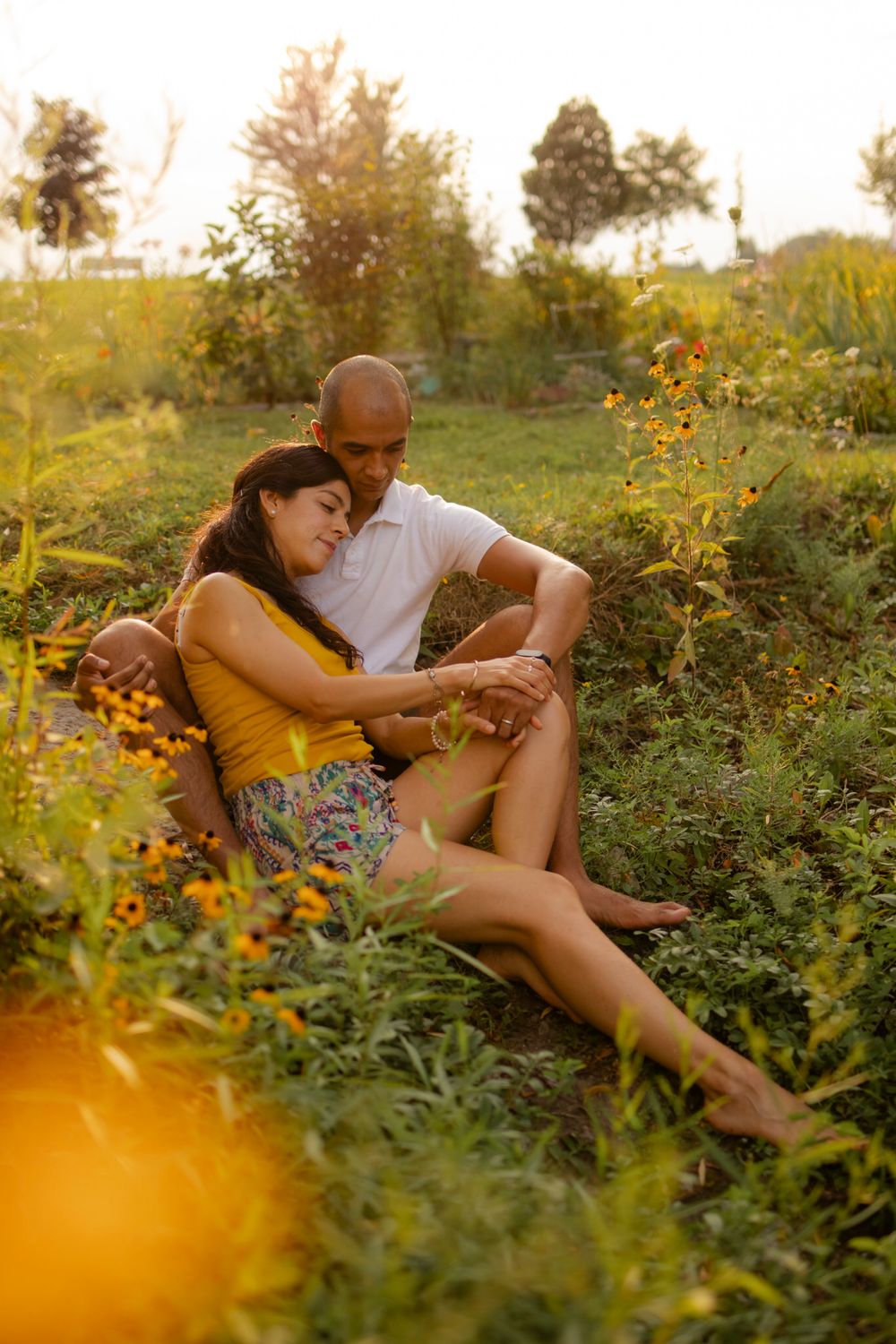 Couple sits among yellow flowers at sunset/golden hour. She sits in his lap and lays head on his shoulder. Kingston, ON, CA.