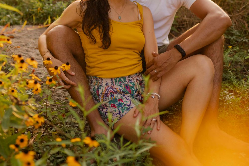 Couple sits among yellow flowers at sunset/golden hour. Detail: hands as she sits in his lap on the shore at Lake Ontario, Kingston, ON, CA.