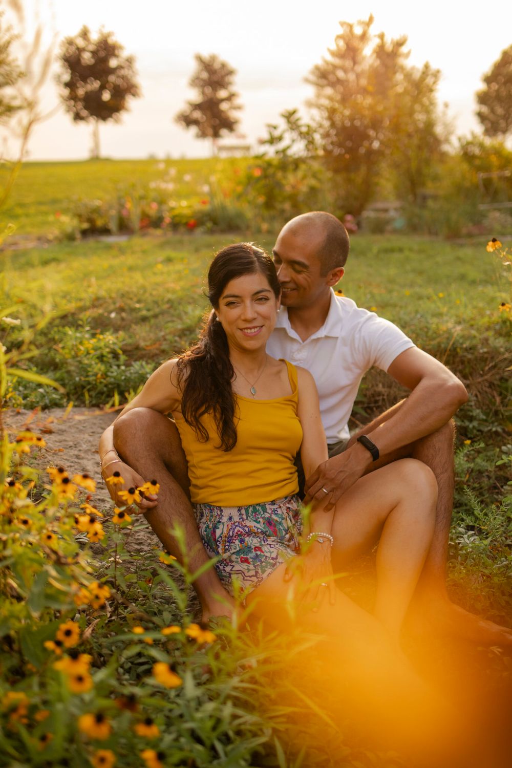 Couple sits among yellow flowers at sunset/golden hour. She sits in his lap on the shore at Lake Ontario, Kingston, ON, CA.