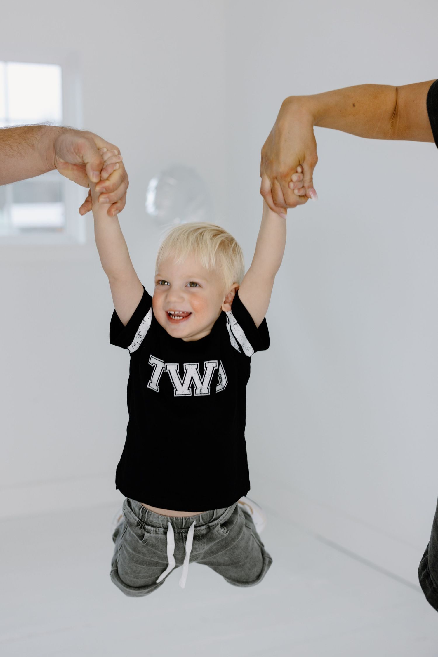 A joyful child in a black t-shirt with TWS print holding hands with adults while standing in a bright white room.