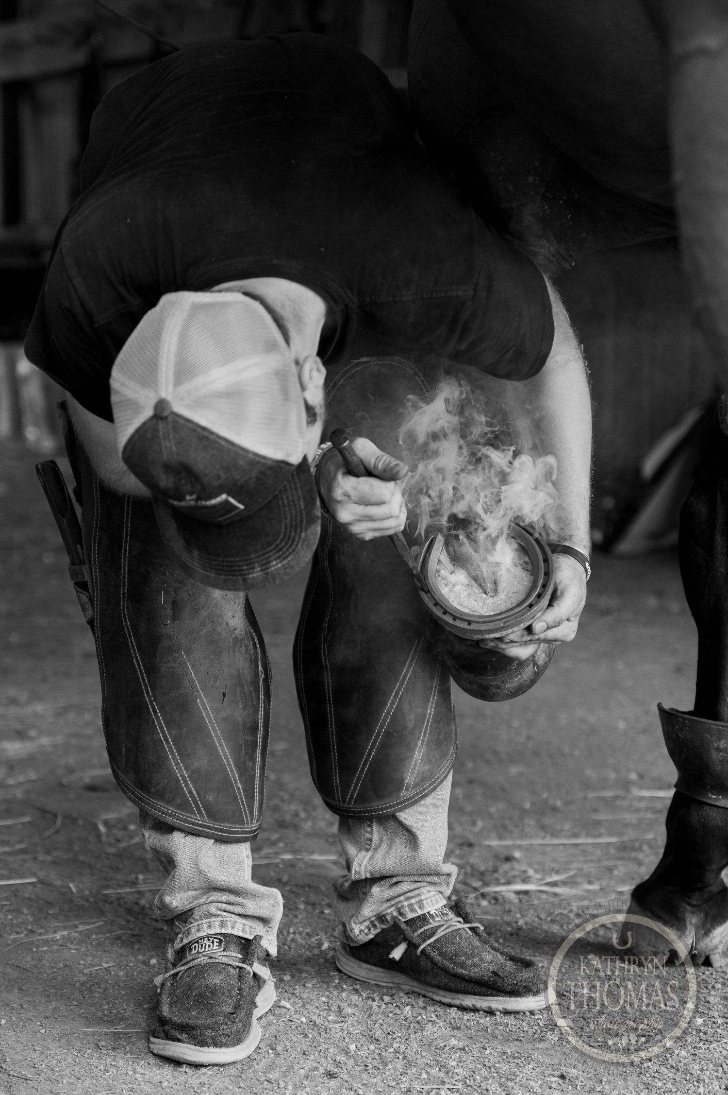 A black and white closeup of worn boots and jeans with smoke rising from a farrier's work.