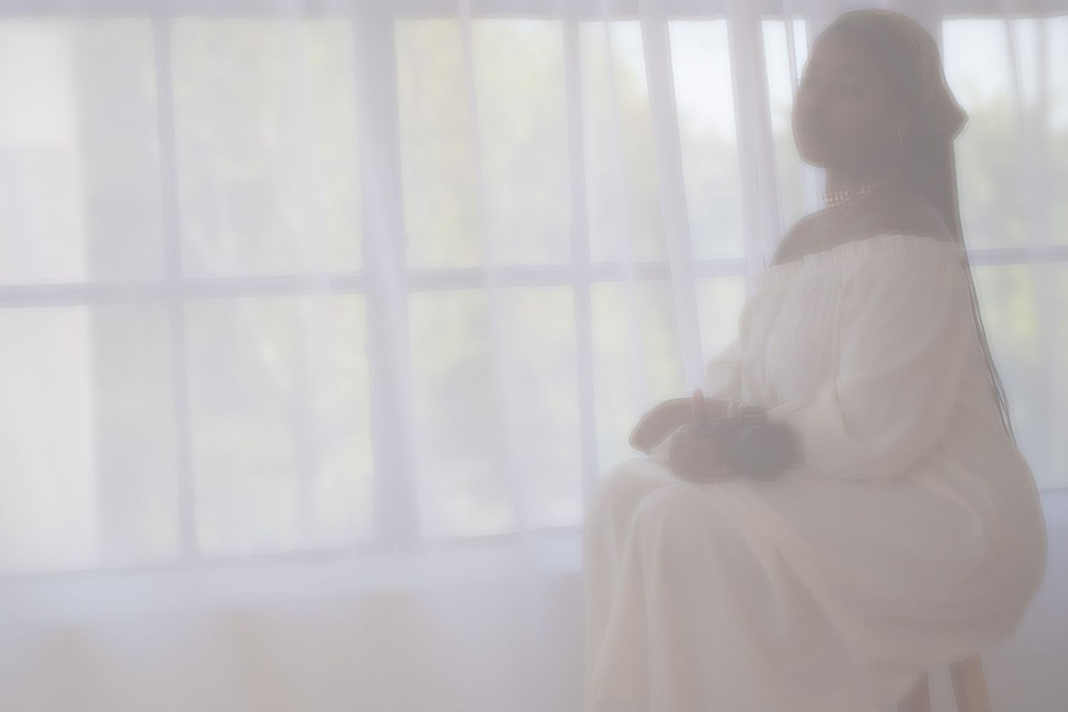 A blurred silhouette in white robes stands near a sheer curtained window creating an ethereal atmosphere.