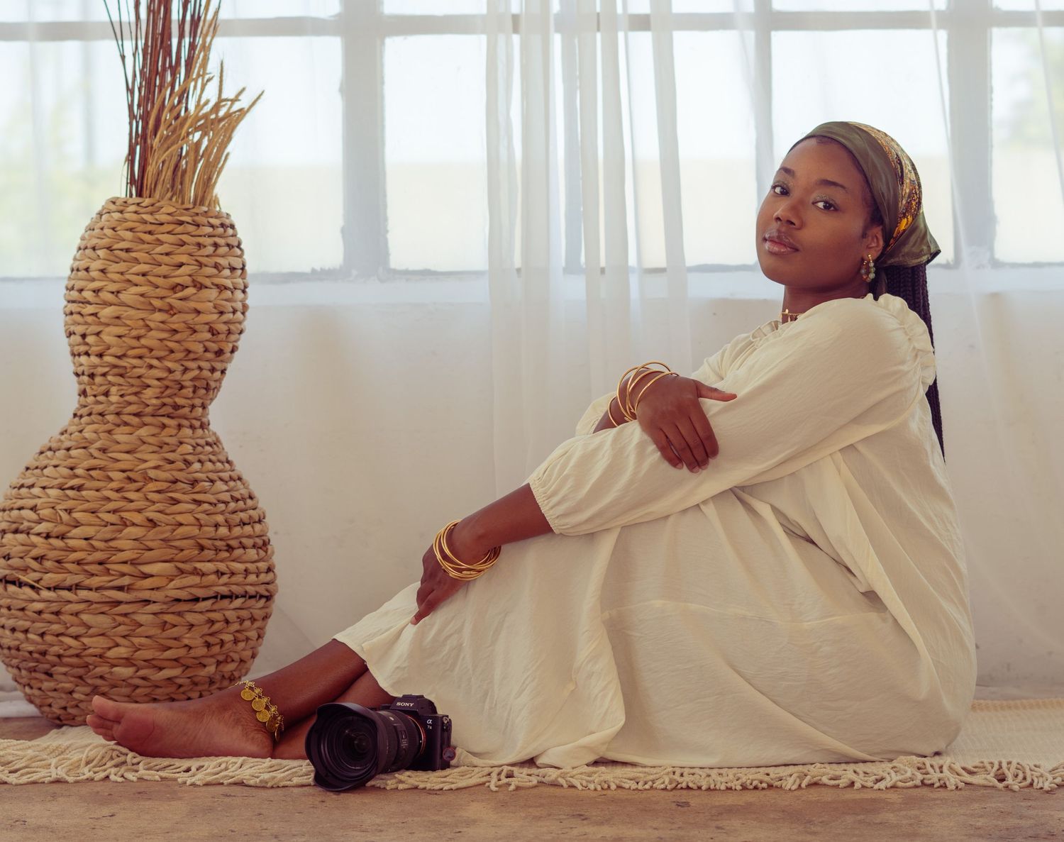 A person in a flowing white dress relaxes against a wall beside a woven decorative vase in a bright sunlit room.