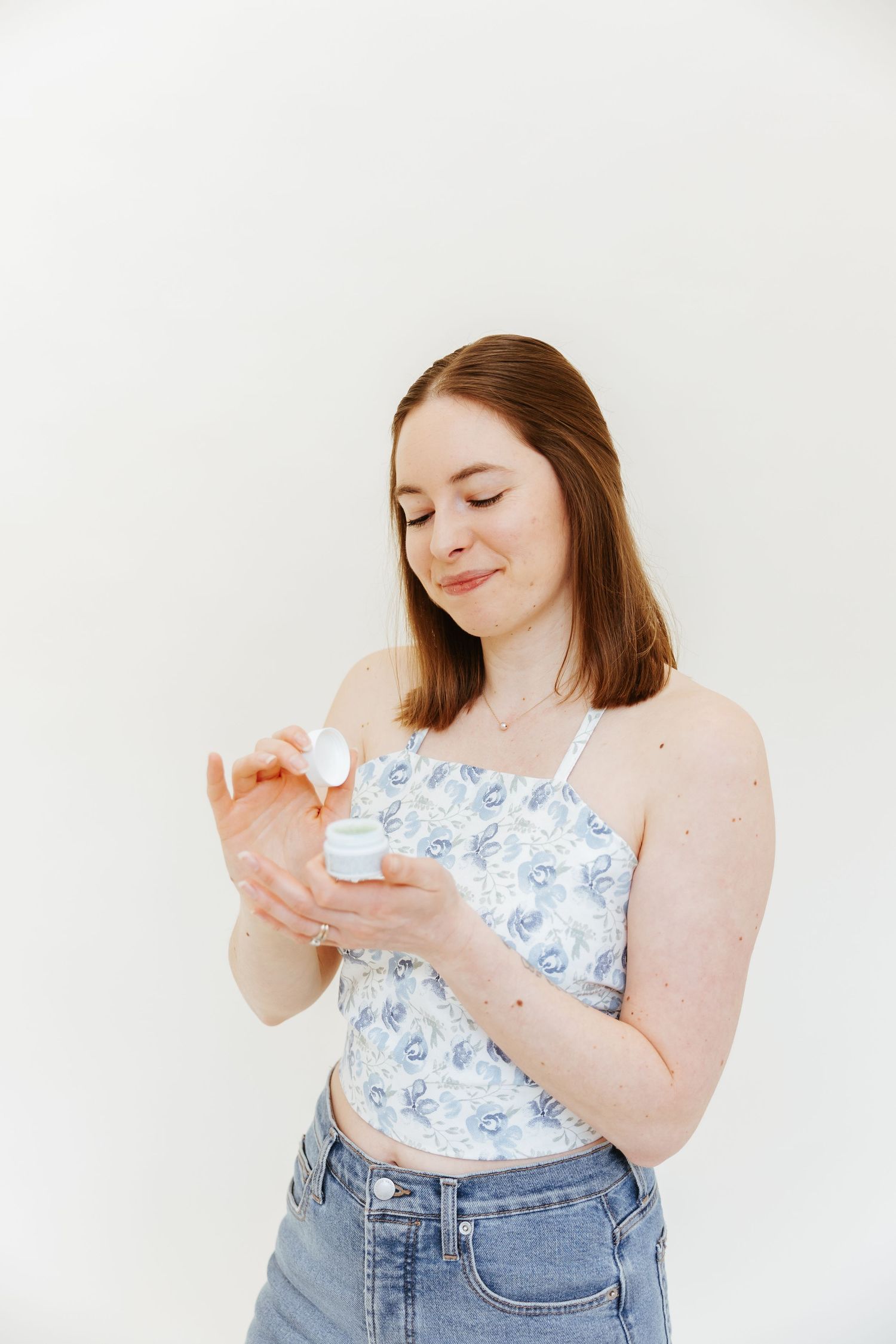 Woman in floral crop top and jeans gazes down while holding skincare product against white background.