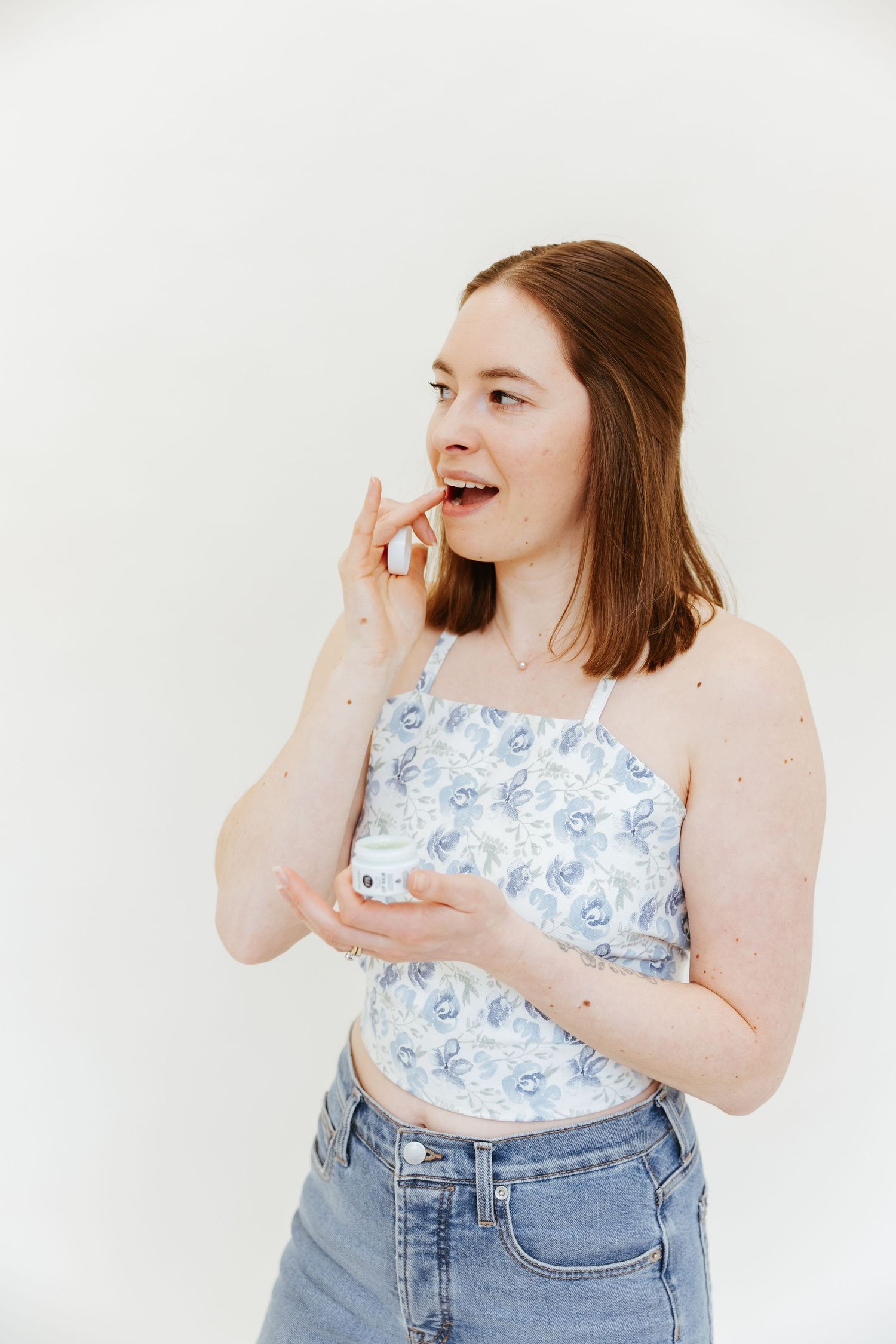 Person in floral crop top and jeans holding CBD product against white wall.