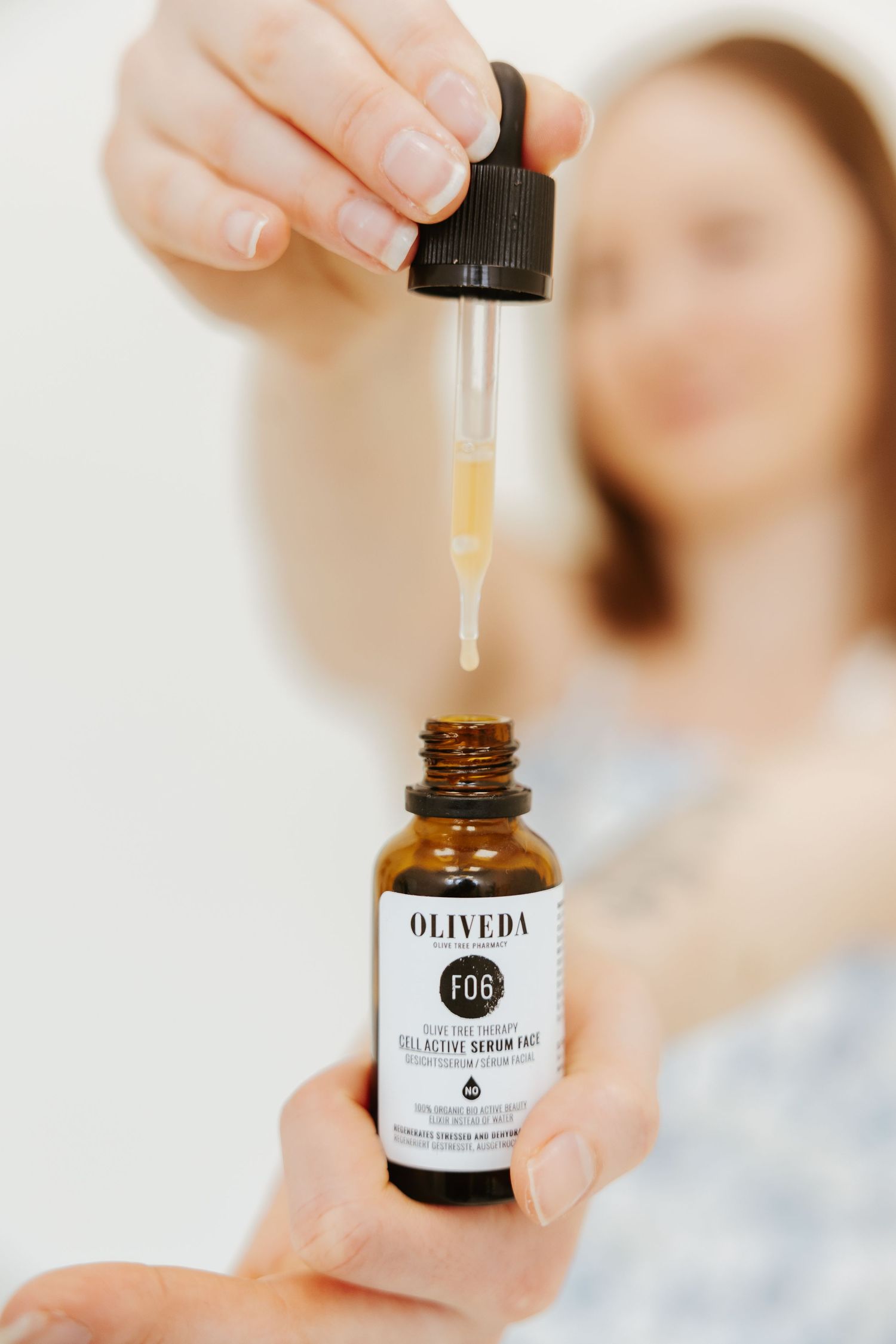 Close-up of hands dispensing dropper from amber glass bottle of Oliveda skincare product.