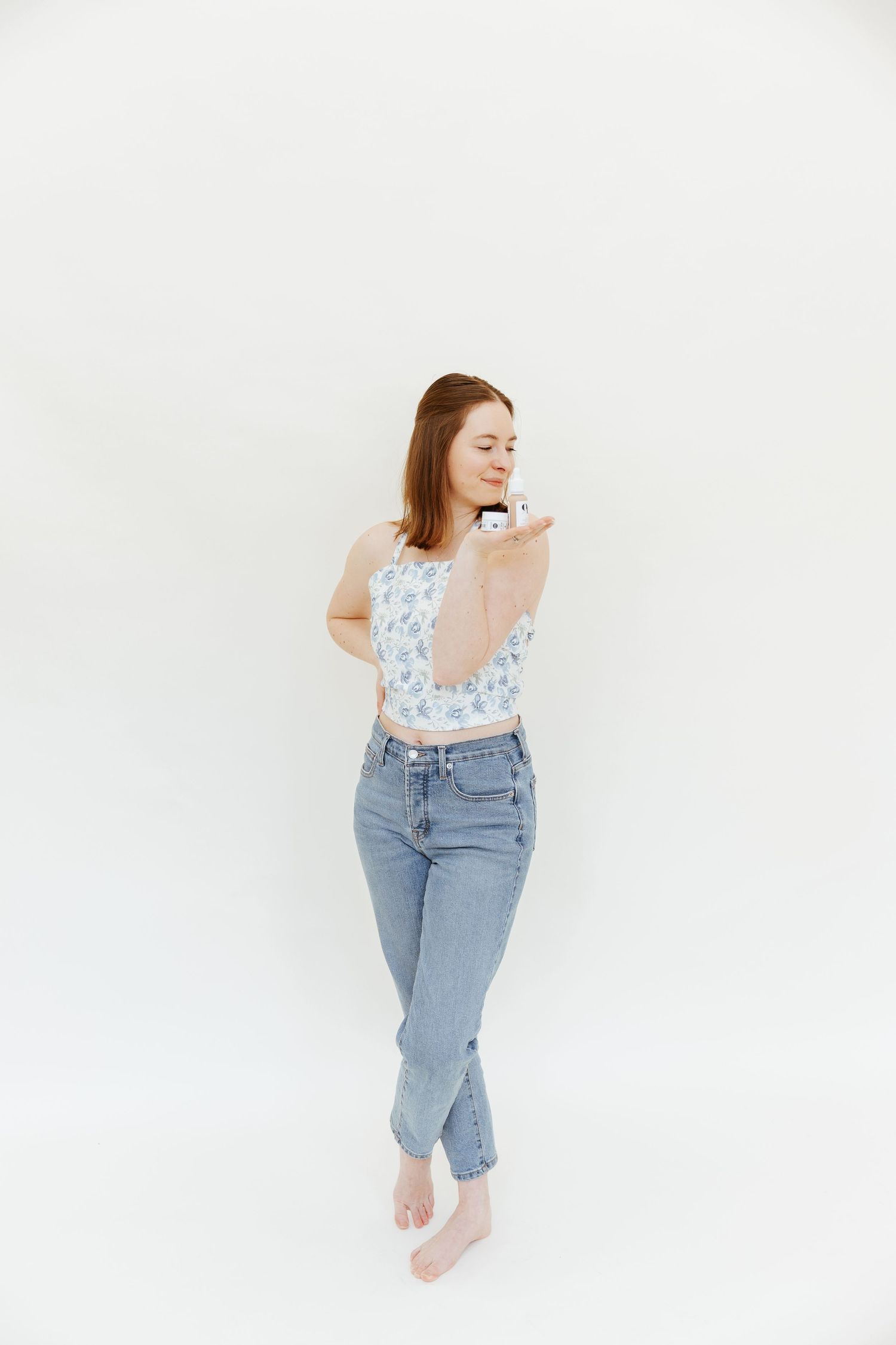 Woman in floral top and jeans poses against white wall with bare feet and crossed legs.