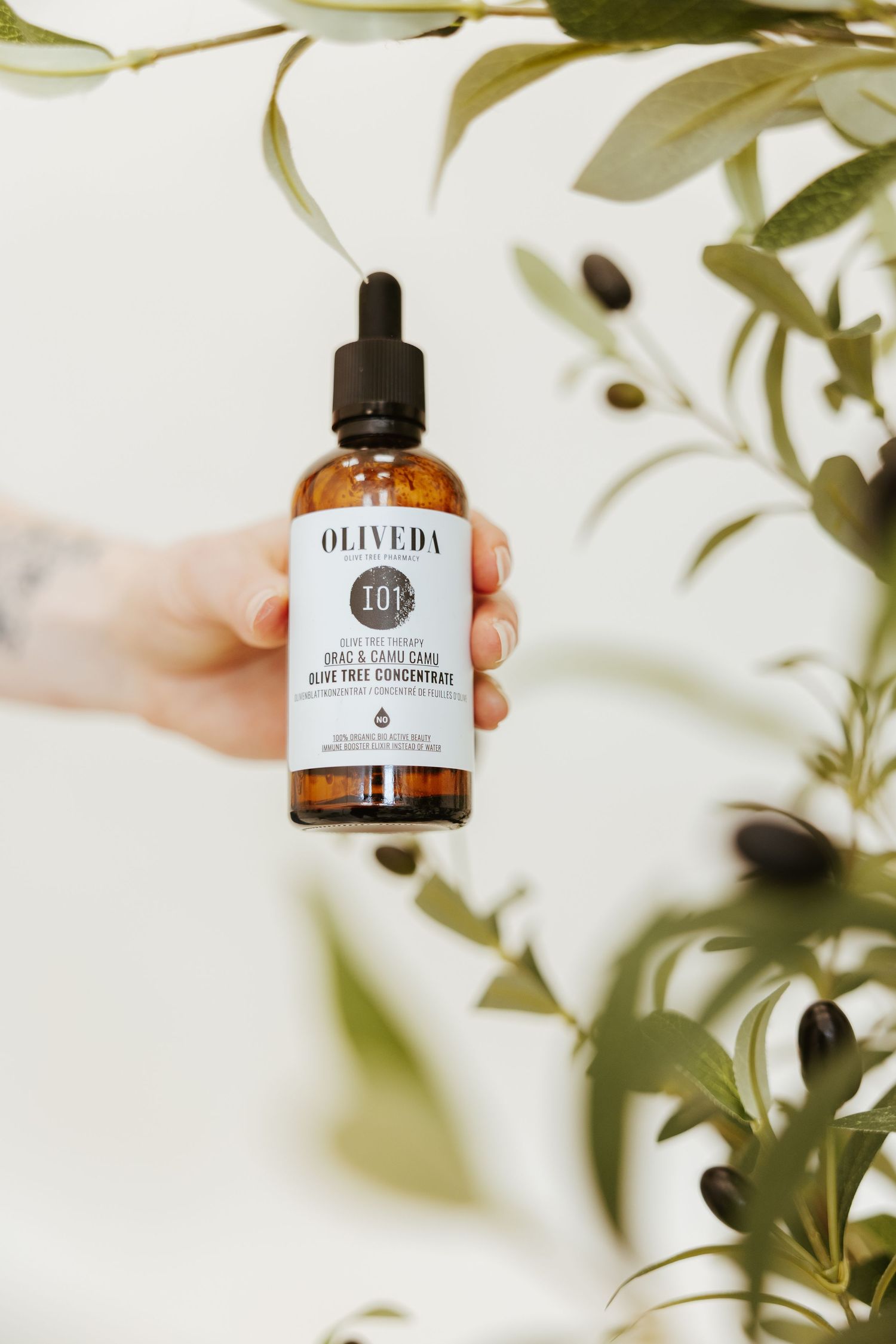 Amber glass bottle of Oliveda skincare product displayed with green foliage against white background.