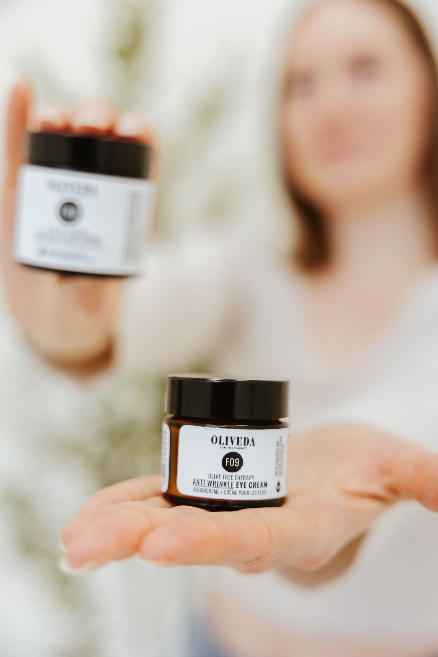 Two black jars of Oliveda skincare products held against blurred background.