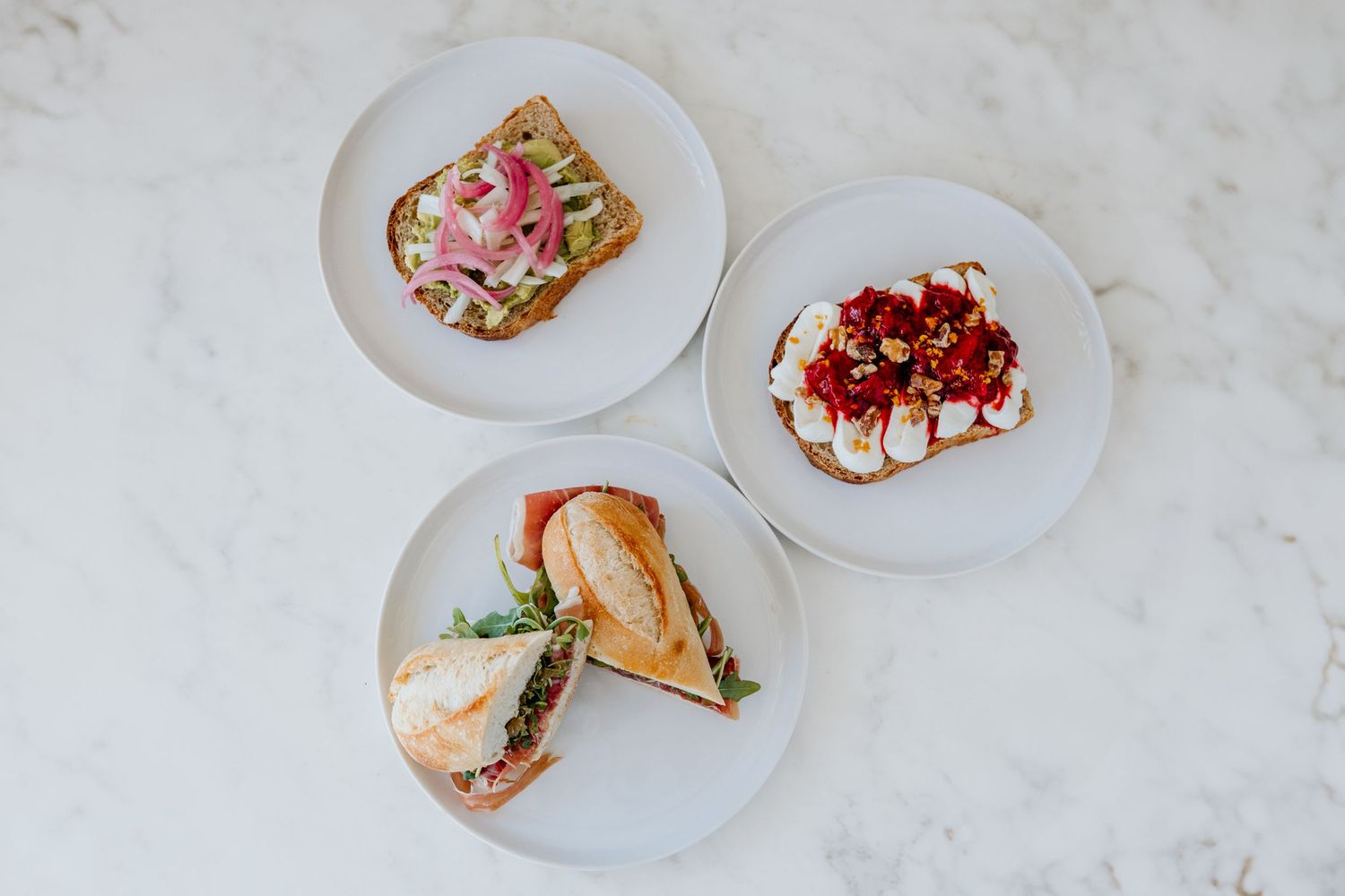 Three plates of artisanal toast topped with various ingredients including strawberries, pink onions, and herbs on a white surface.