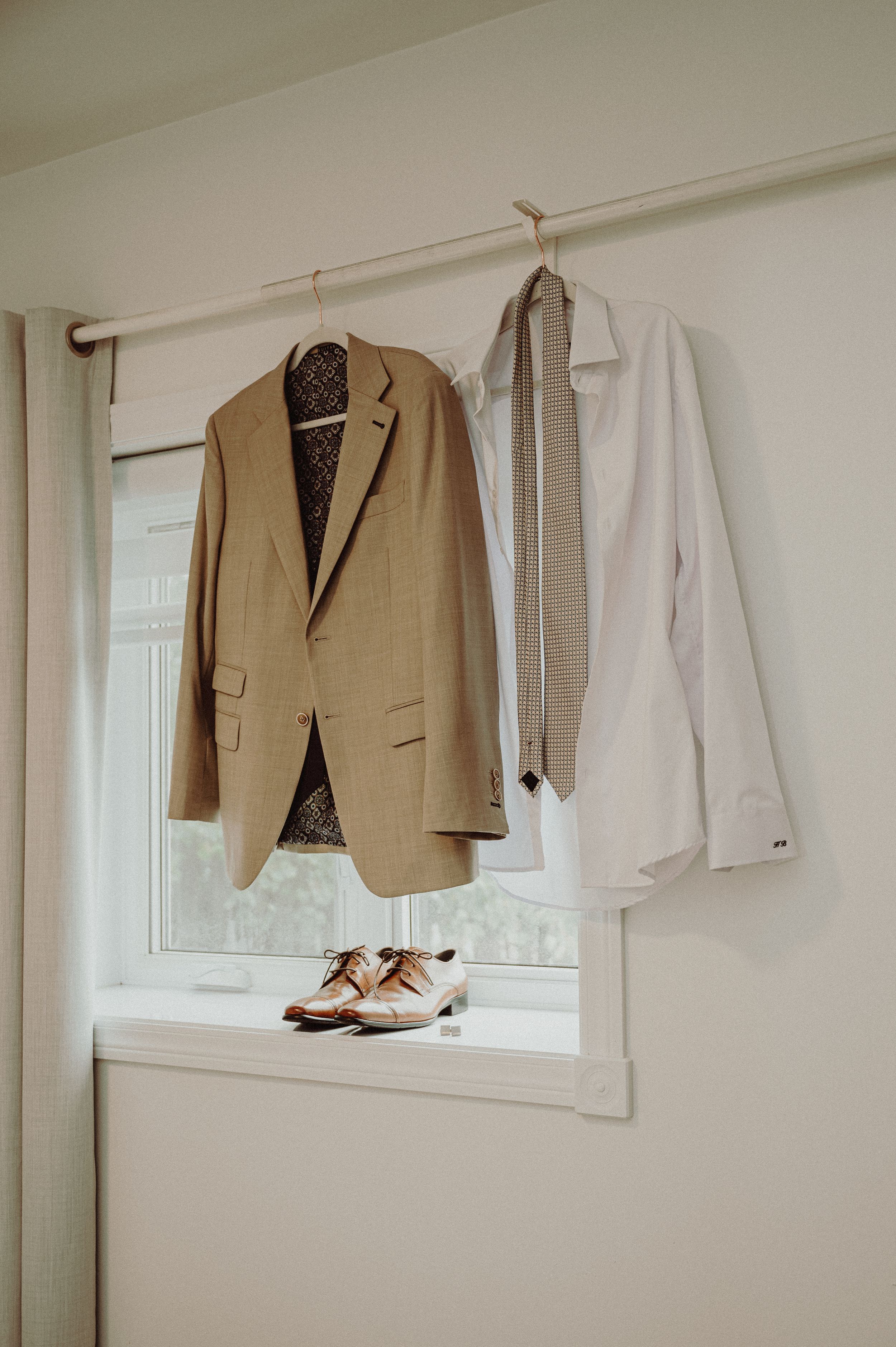 Wedding attire including a tan suit jacket, white shirt, tie and dress shoes hanging by a window.