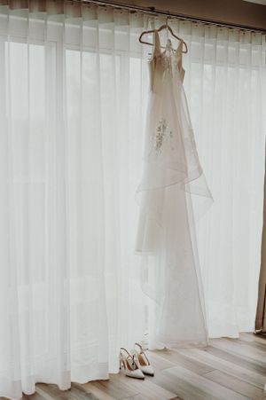 An elegant white wedding dress and heels displayed against sheer curtains in natural light.