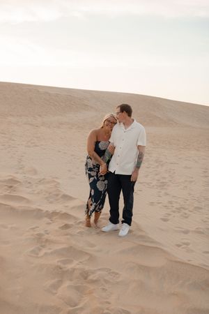 A couple shares romantic moments together while standing on sandy dunes in a desert landscape during golden hour.