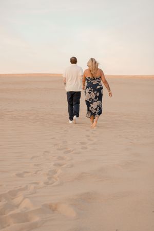 Two people walking together across desert sand dunes at sunset in summertime with footprints trailing behind them.