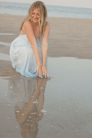 Woman in white dress playing with reflections in wet sand at the beach during sunset.