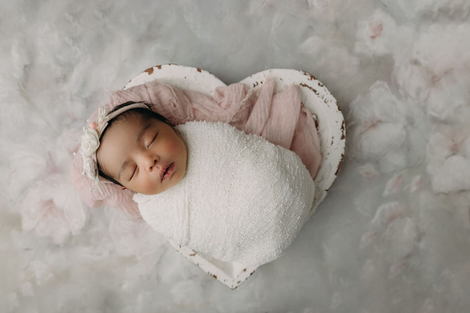 A newborn baby girl swaddled in white cloth lies peacefully on a heart backdrop with pink accents for Tampa Newborn photography session