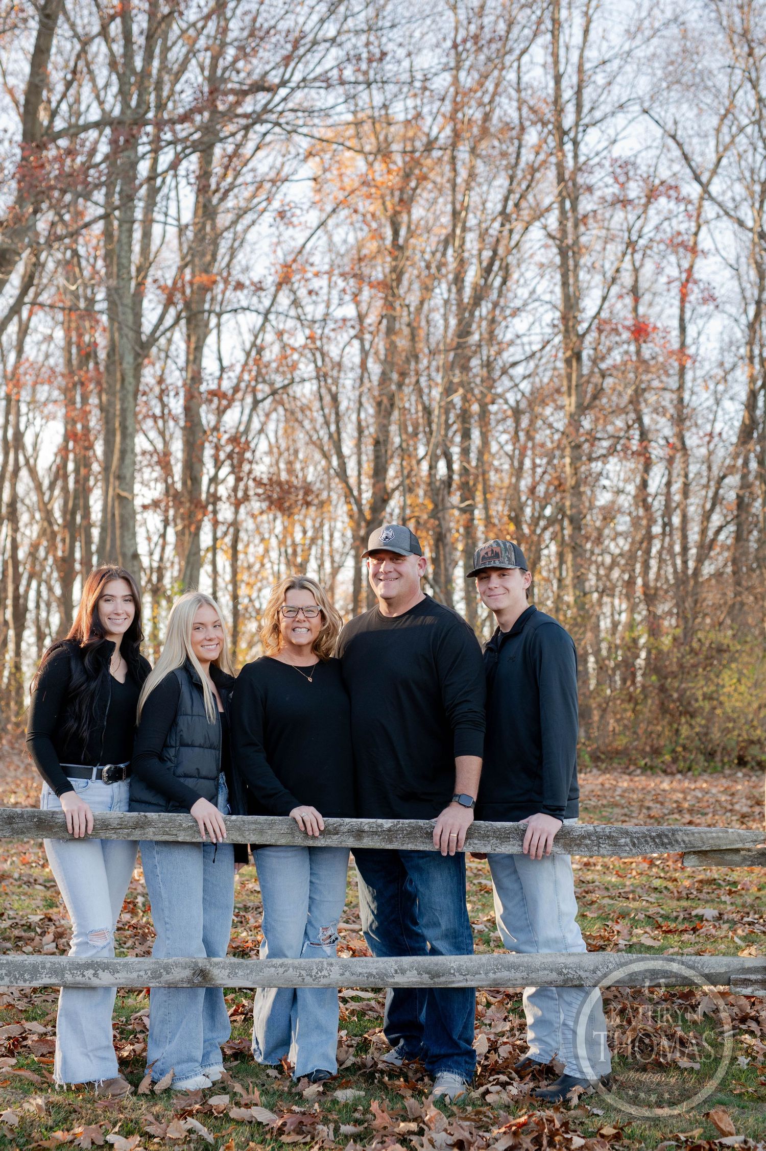 Five people in casual attire stand together at a wooden fence with bare trees in the background.
