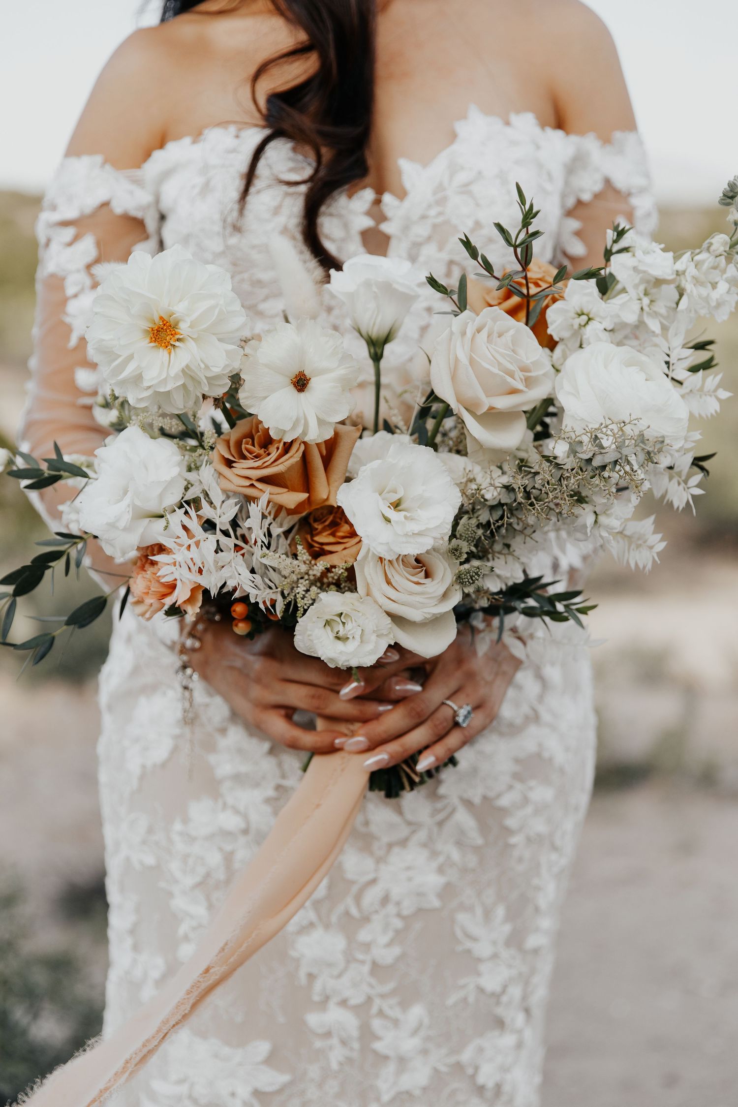 A wedding bouquet of white and beige roses with greenery held against a lace wedding dress.