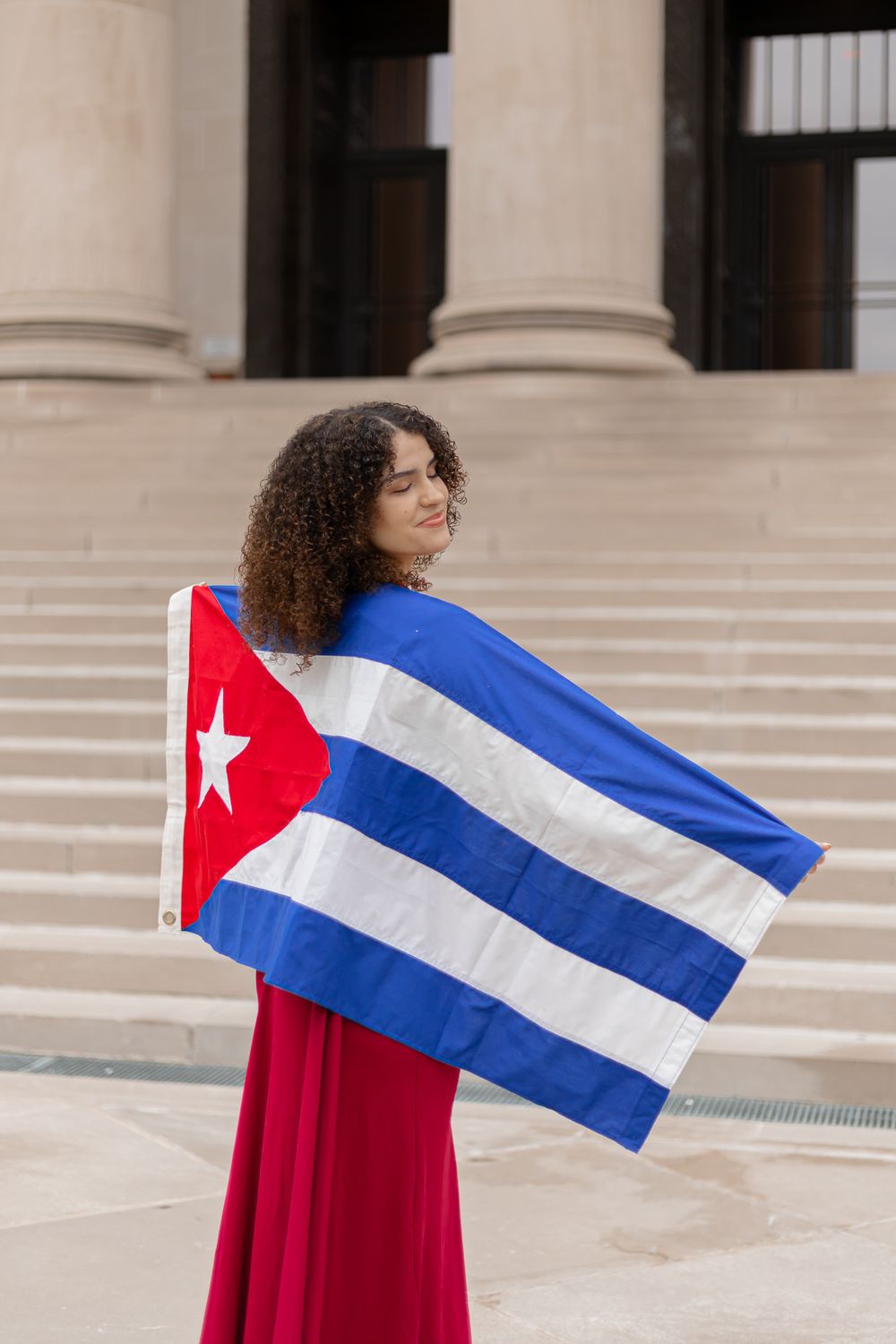 Person in red dress holding Cuban flag in front of classical building columns.