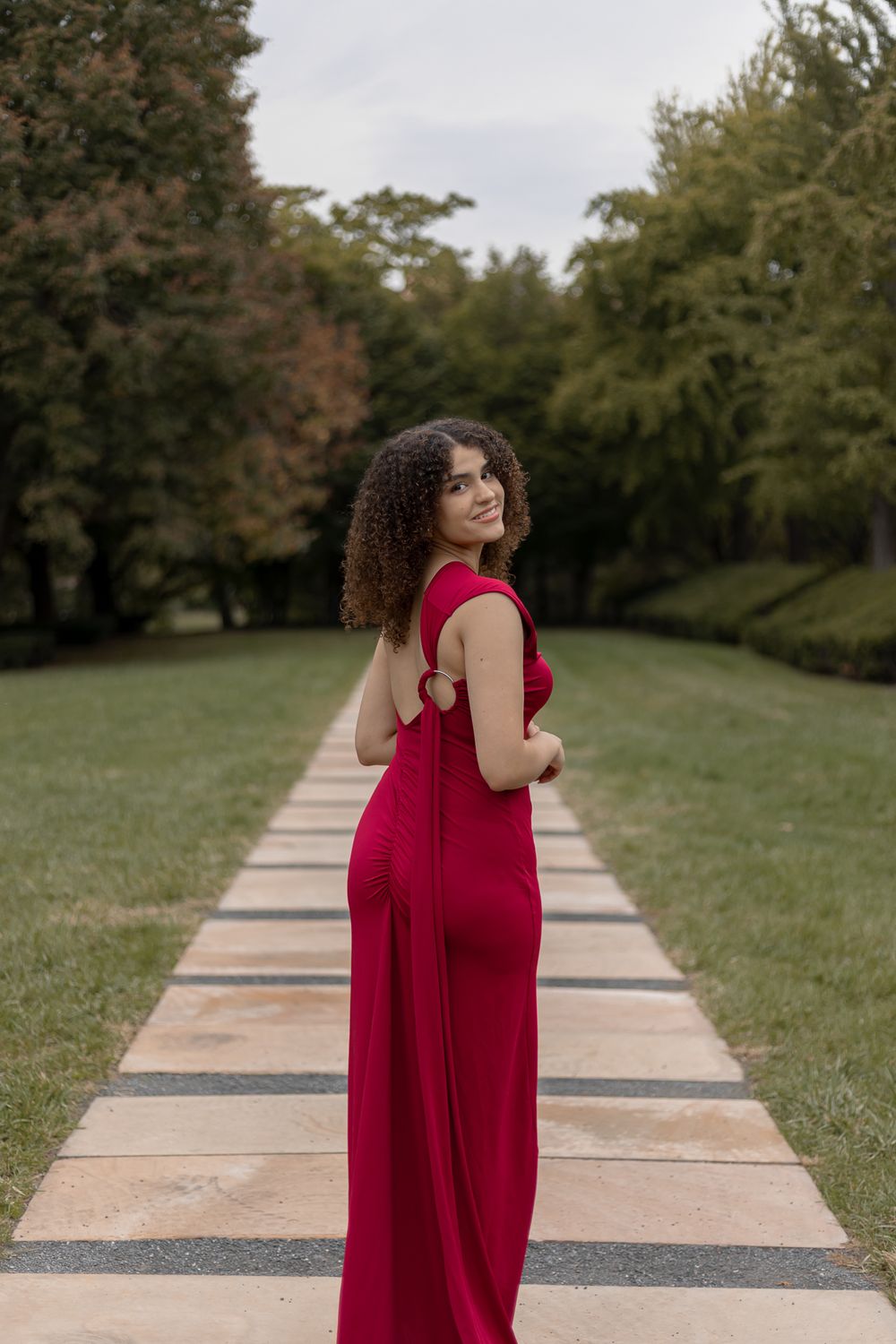 Person in red formal dress poses on garden path surrounded by trees.