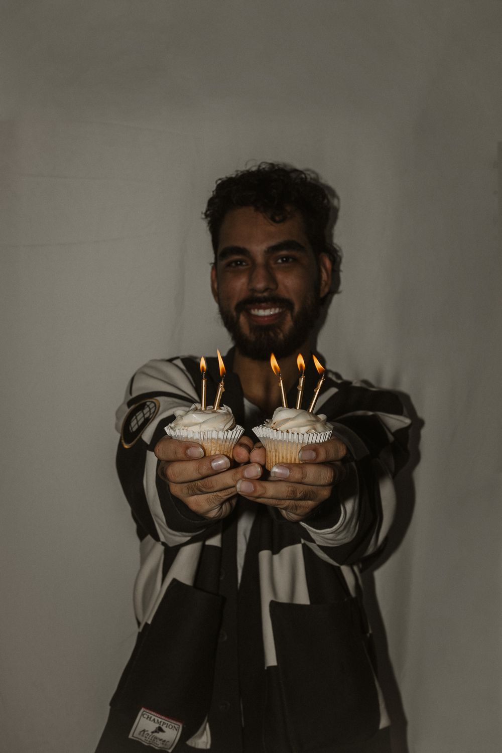 Person holding two cupcakes with lit birthday candles and smiling in a dark room.