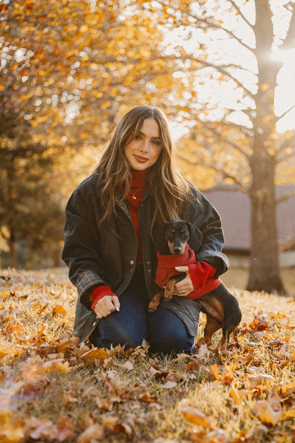 Woman in black jacket kneeling with small dog in red sweater among fallen autumn leaves.