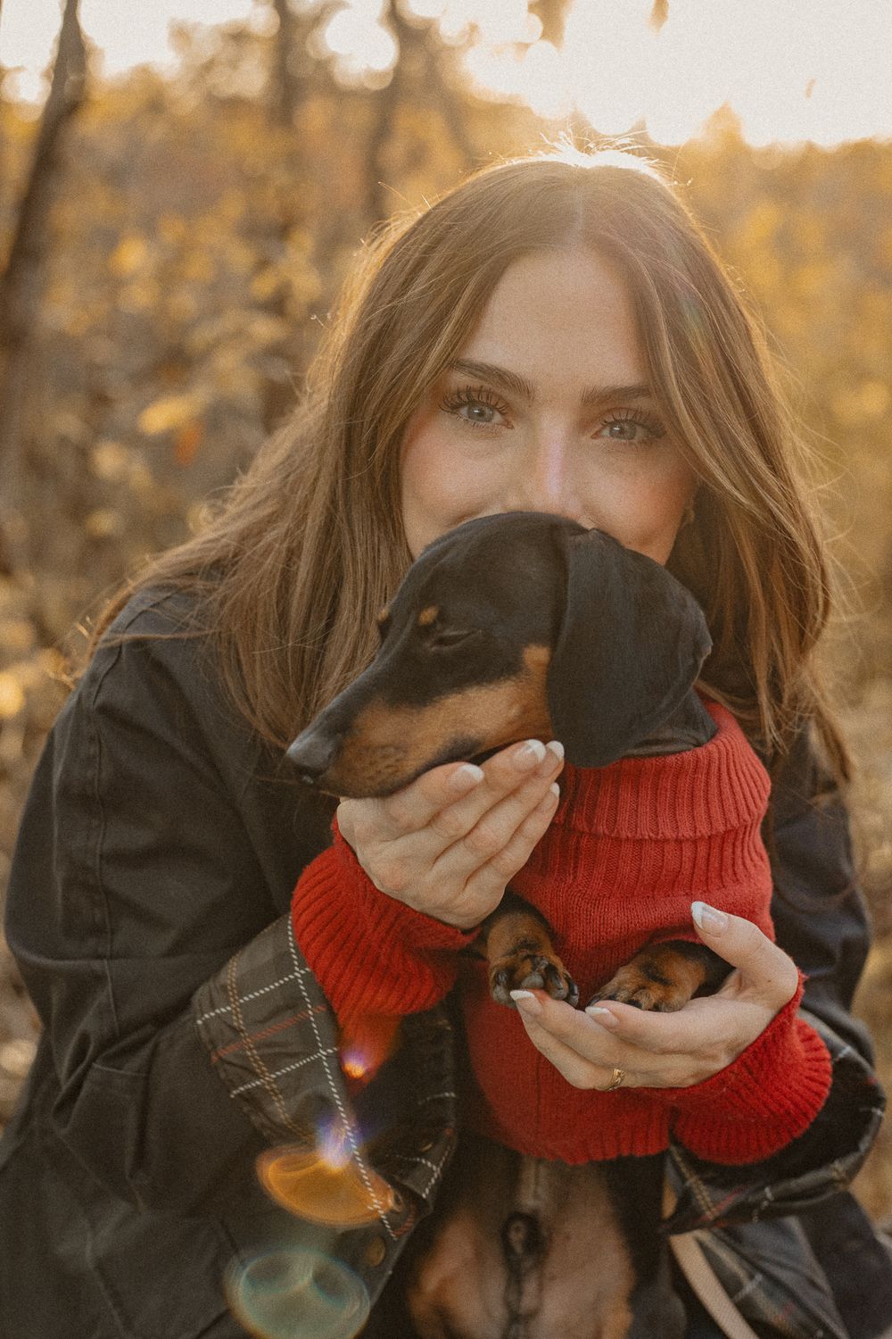 A dachshund puppy wearing a red sweater being held close in an autumn outdoor setting.