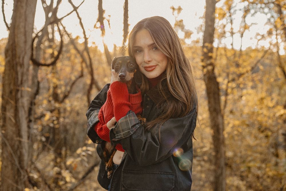 Person holding small dog wearing red sweater in autumn forest setting.