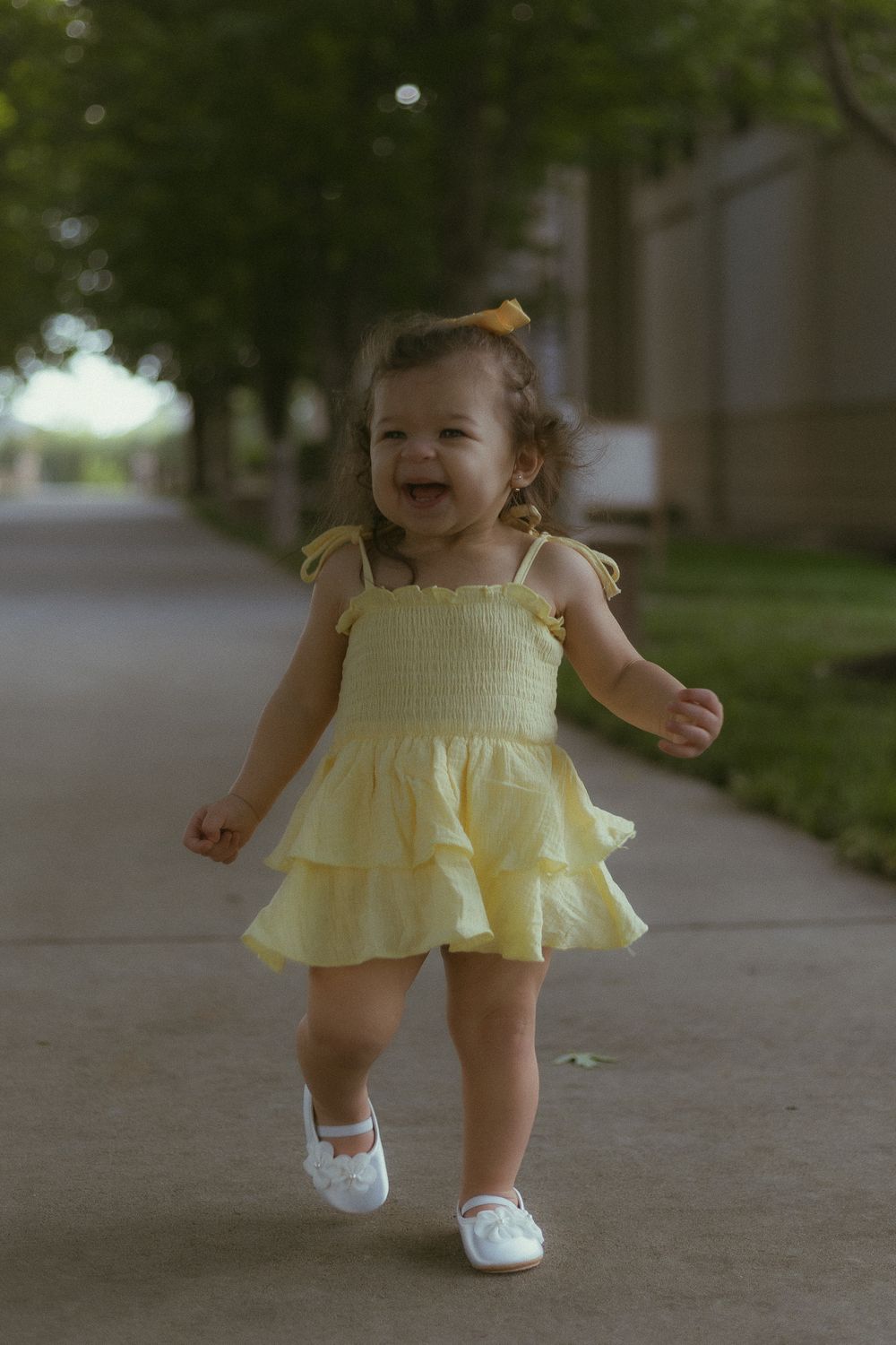 A happy toddler girl in a yellow ruffled dress and white shoes walks on a sidewalk, smiling joyfully.