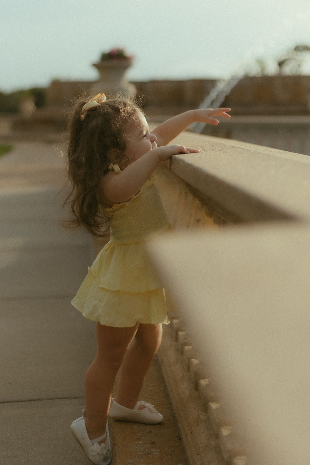 A young girl in a yellow dress reaches toward a fountain wall outdoors in warm golden light.