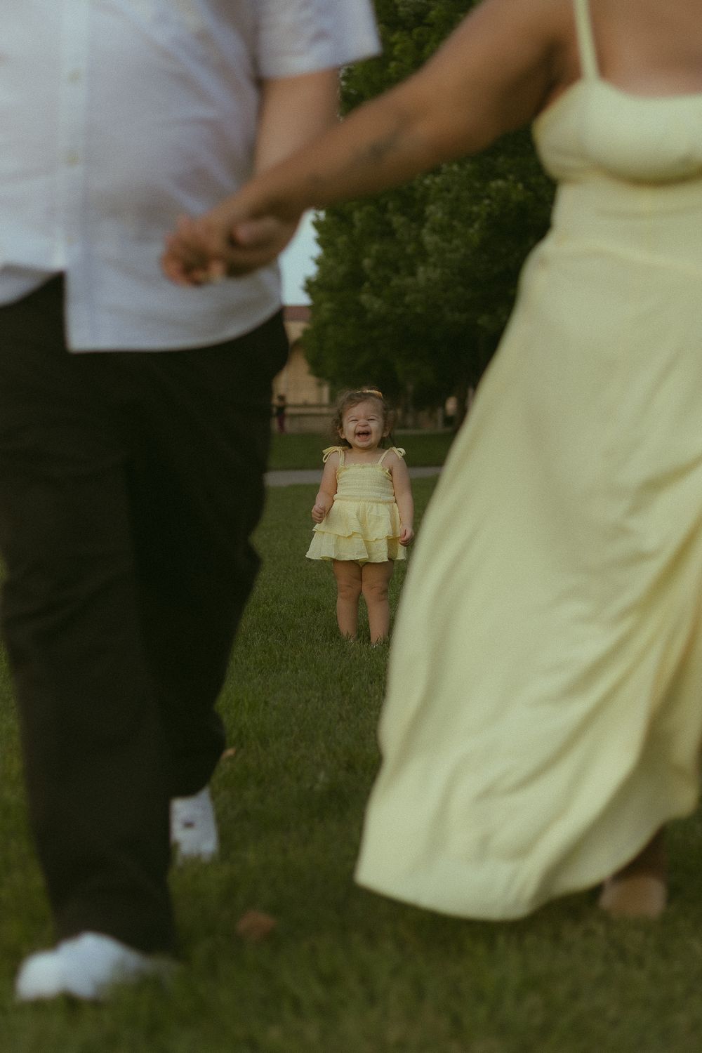 A toddler in a yellow dress stands crying on green grass between two adults during a family photo session outdoors.