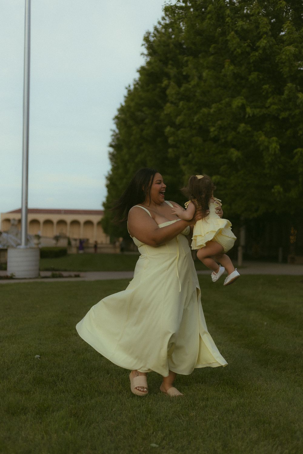 A woman in a yellow maxi dress joyfully lifts a toddler in a green park with trees in the background.