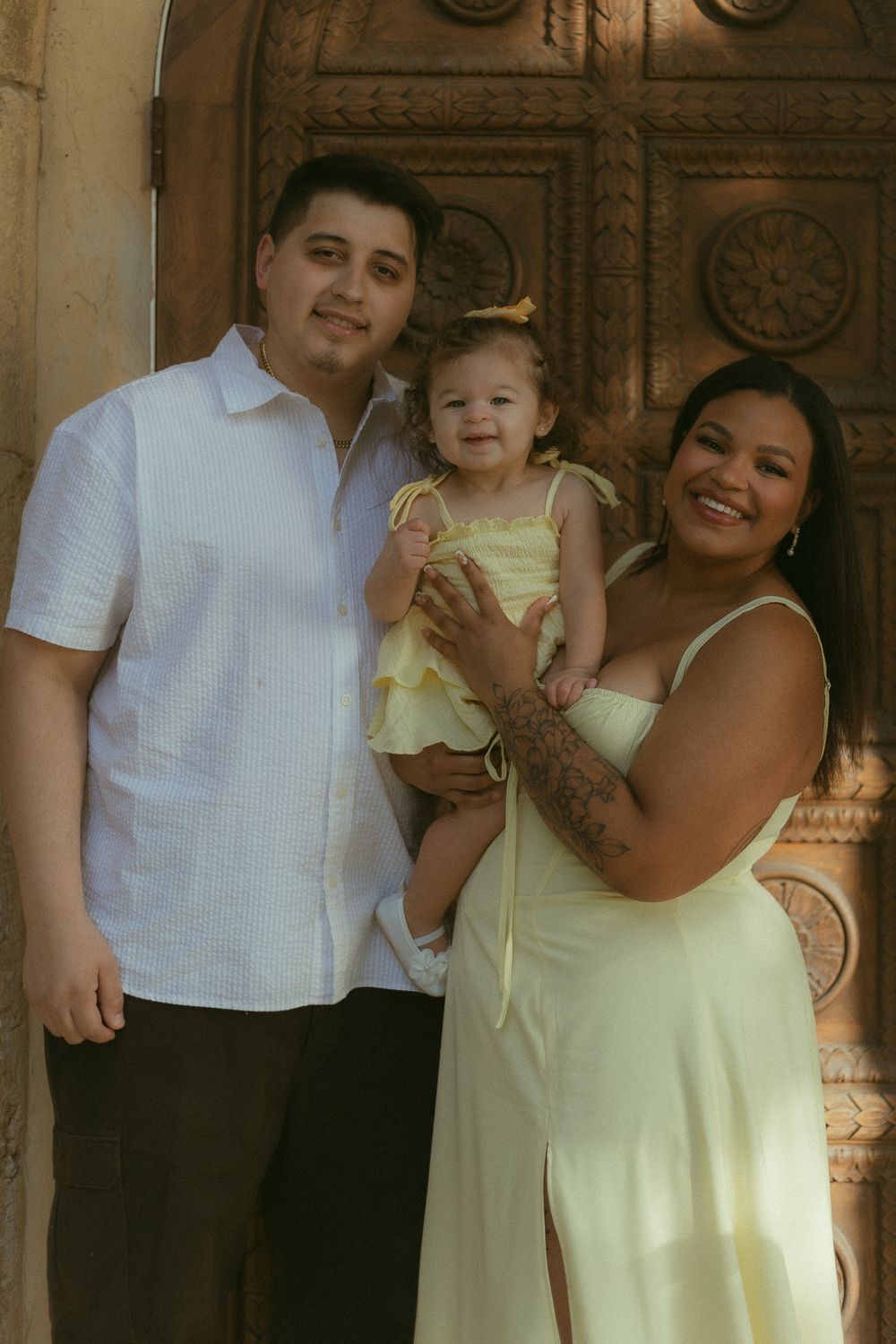 A smiling family of three poses in front of an ornate wooden door, with parents holding their baby girl in a yellow dress.