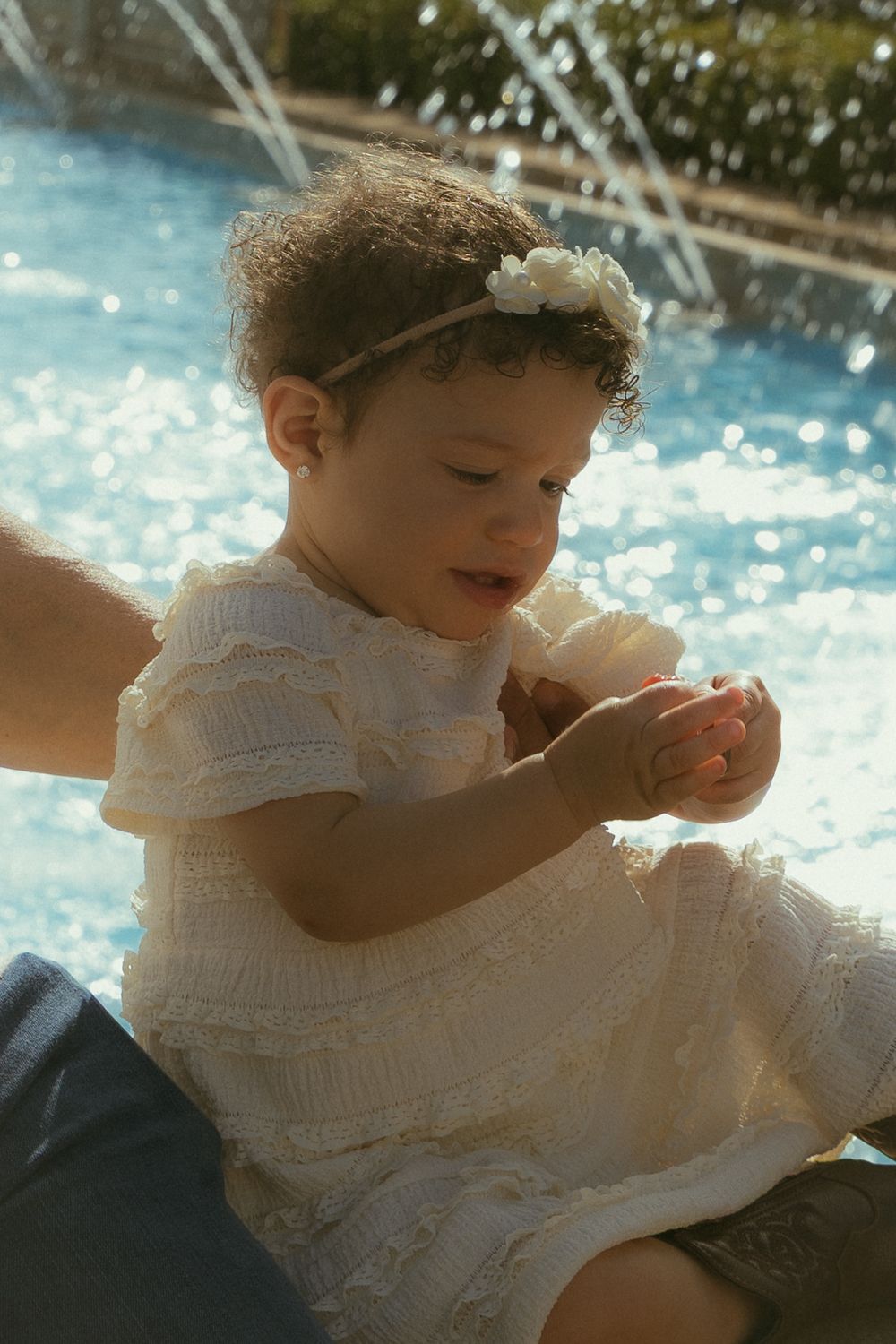 A toddler girl in a white lace dress and floral headband sits near a sunlit fountain.