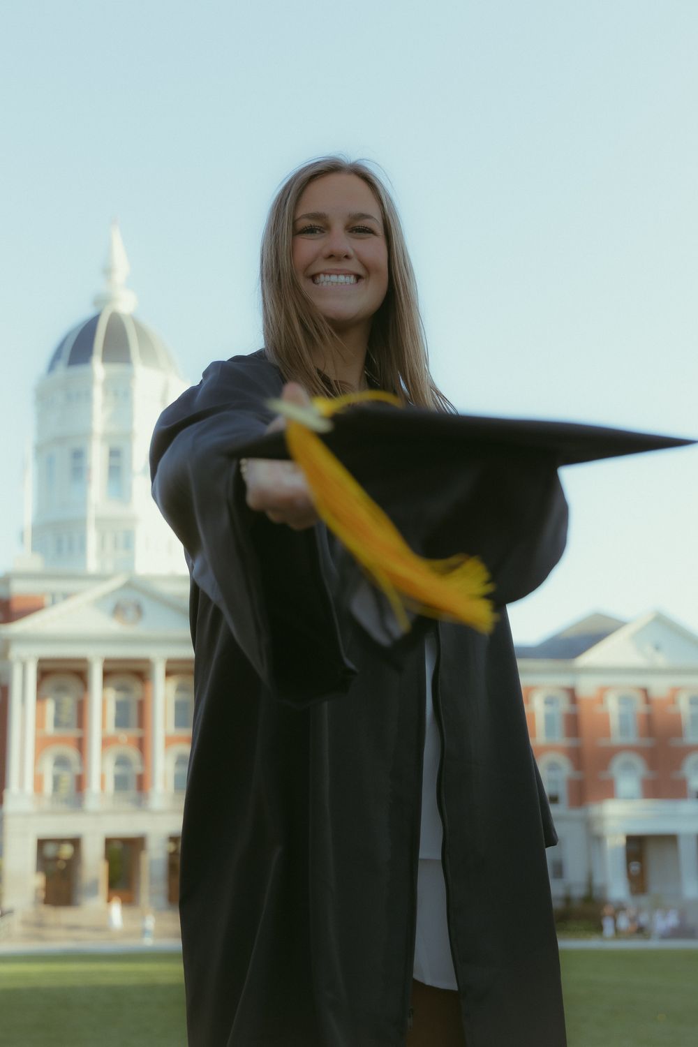 Smiling female graduate in black cap and gown holding mortarboard with yellow tassel outdoors near brick building.