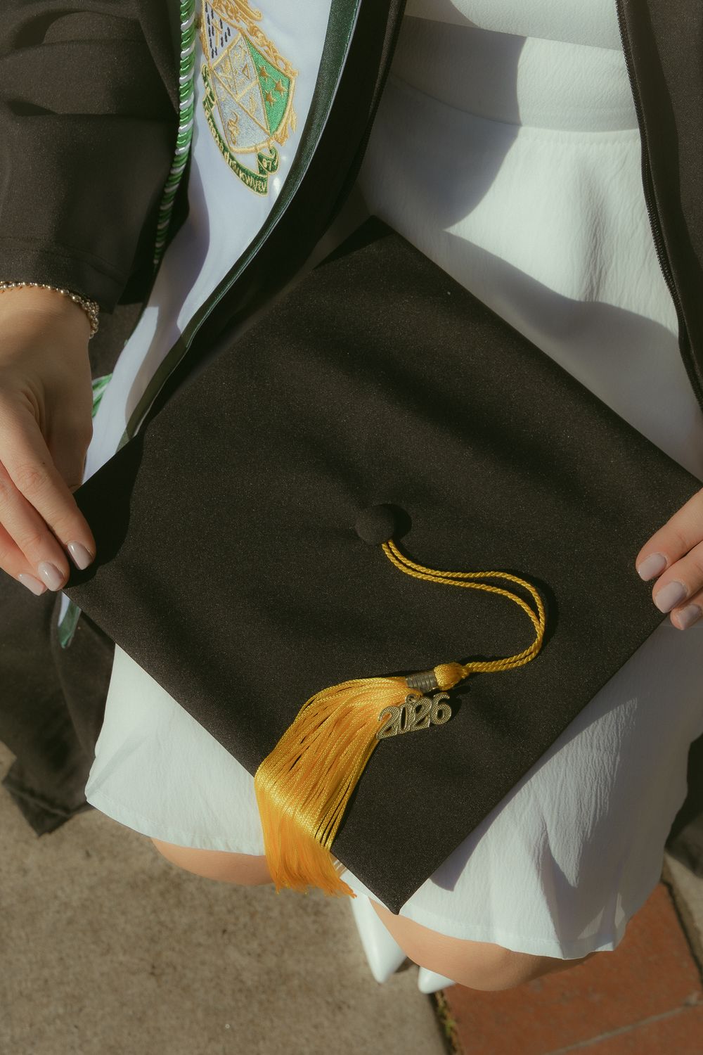 Graduate holding a black mortarboard cap with gold tassel and 2026 charm, wearing academic regalia with green honor sash.