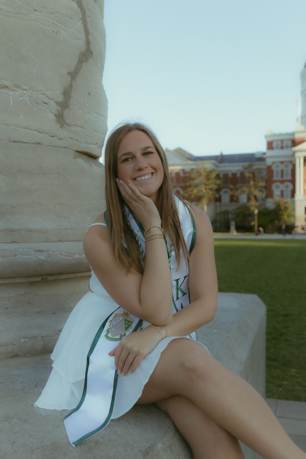 Smiling young woman in white dress and Greek sorority sash sits on stone steps of college campus building.