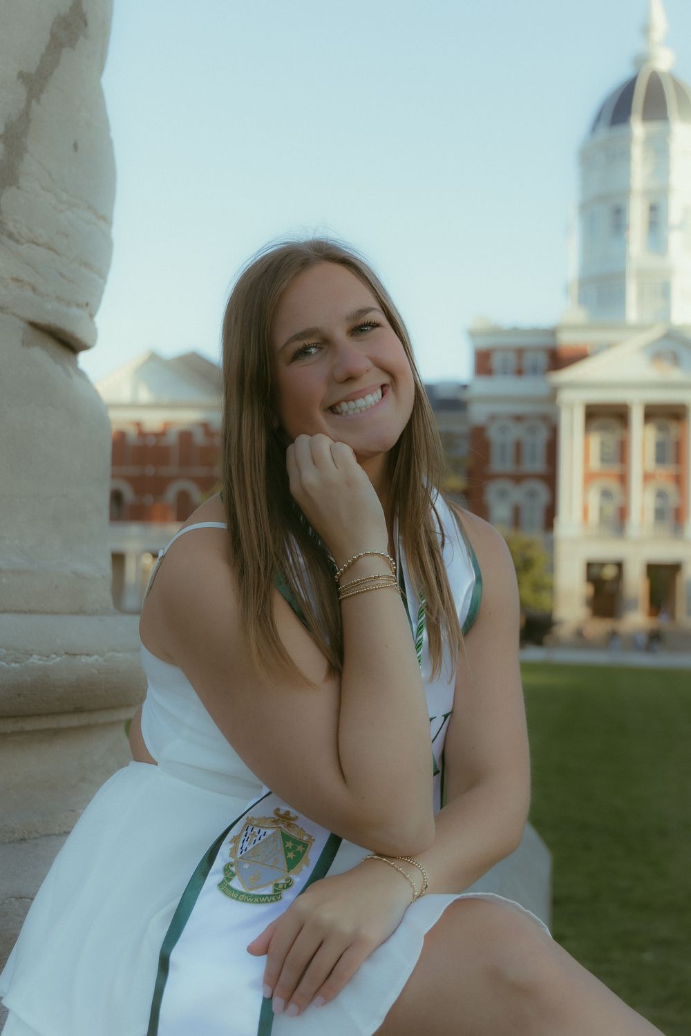 Smiling young woman in white dress sitting outdoors near classic architecture on a sunny day.