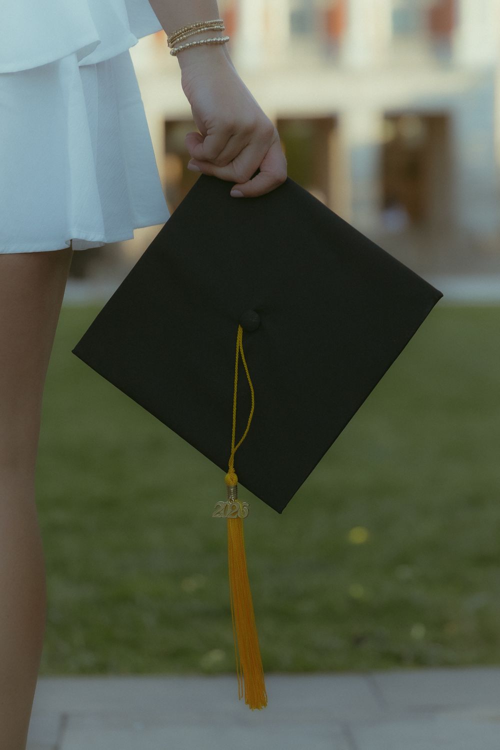 A graduate in a white dress holds a black mortarboard cap with a gold tassel outdoors.