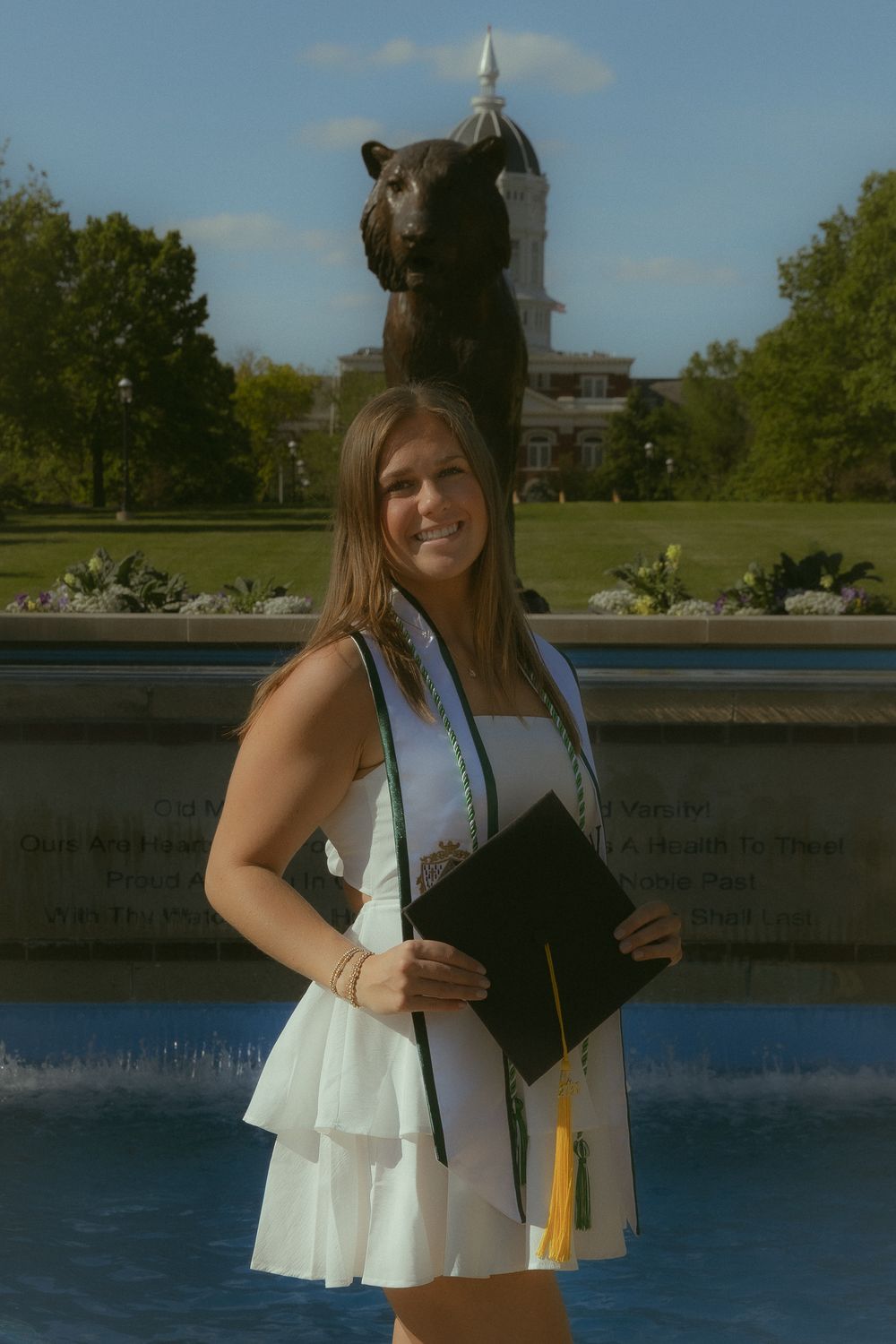 A smiling graduate in a white dress holds her cap near a bear statue fountain on a sunny college campus.