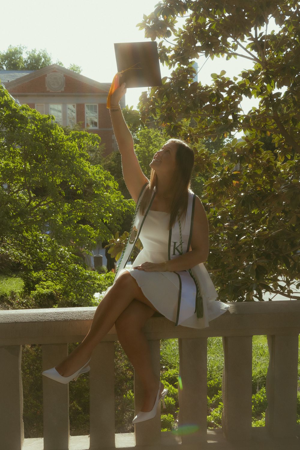 A young woman in a white dress holds her diploma aloft while seated on a railing at her college graduation.