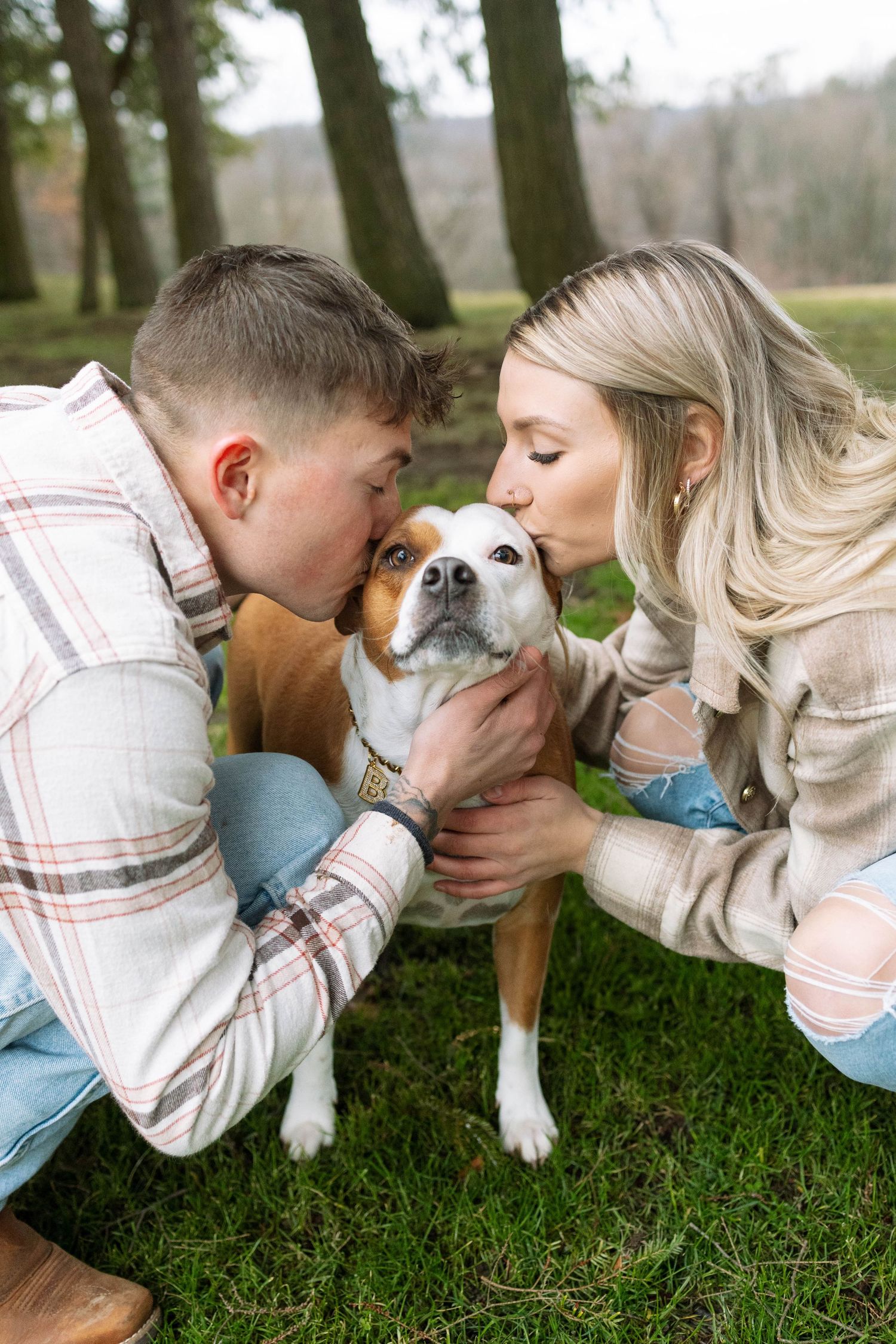 A couple giving love and affection to their brown and white dog while sitting on grass in a park.