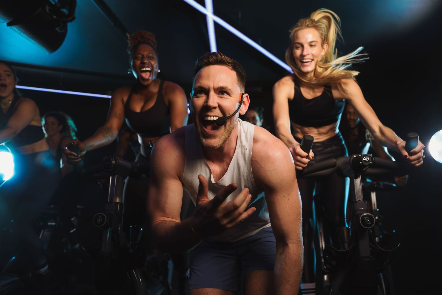 Energetic group exercise class with people riding spin bikes in a dark room with blue lighting.