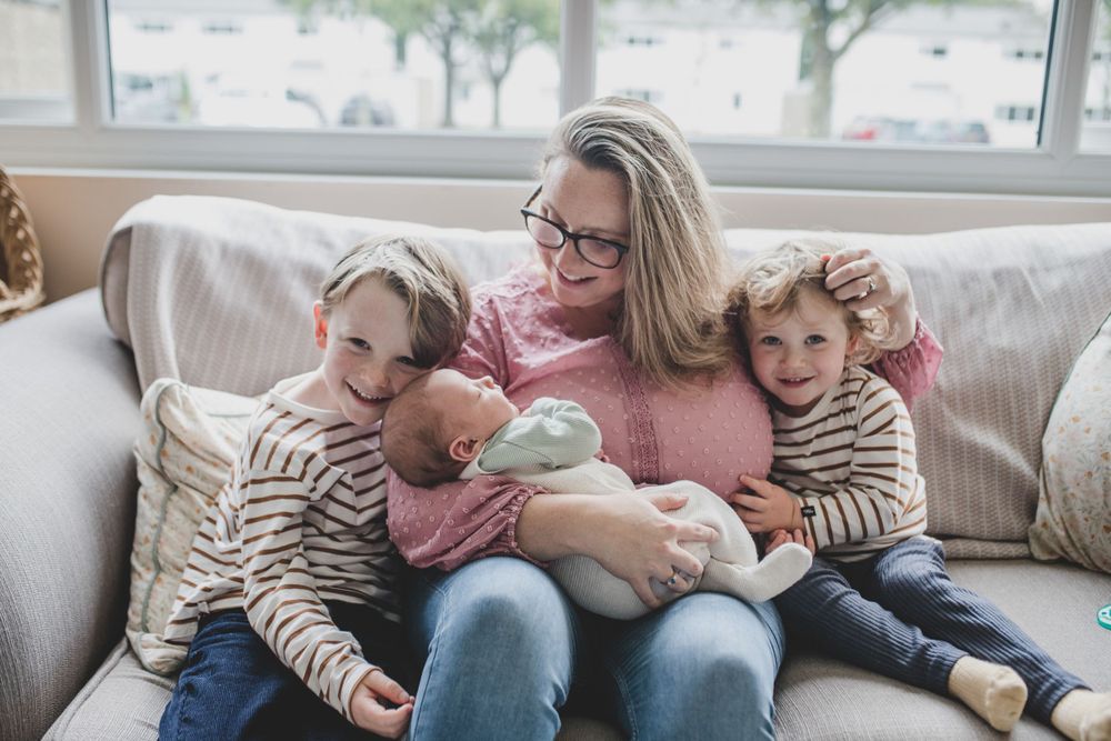 A family cuddles together on a gray couch while bottle feeding a baby as natural light streams through the window.