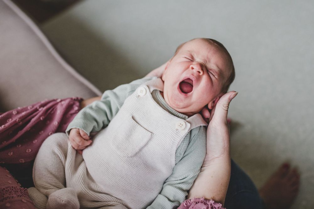 A tired baby yawns while lying in a white and grey outfit against a burgundy blanket.