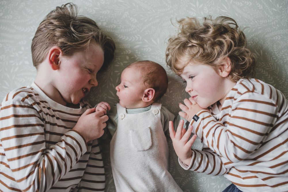 Three siblings in striped shirts lie together on a gray blanket with a small infant in center wearing white.