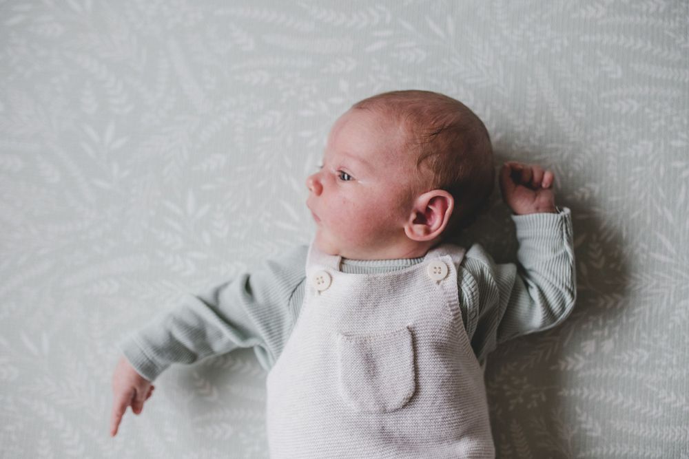 A small infant wearing a light gray onesie lies on white patterned bedding while gazing to the side.