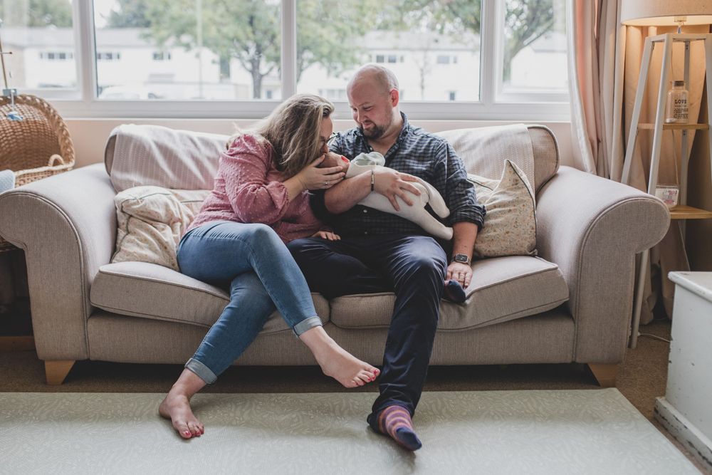 A couple sits together on a cream-colored couch relaxing with their small white dog in a bright living room.