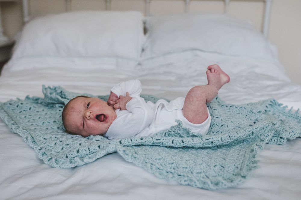 Baby lying on a mint green blanket on white bedding while stretching and yawning.