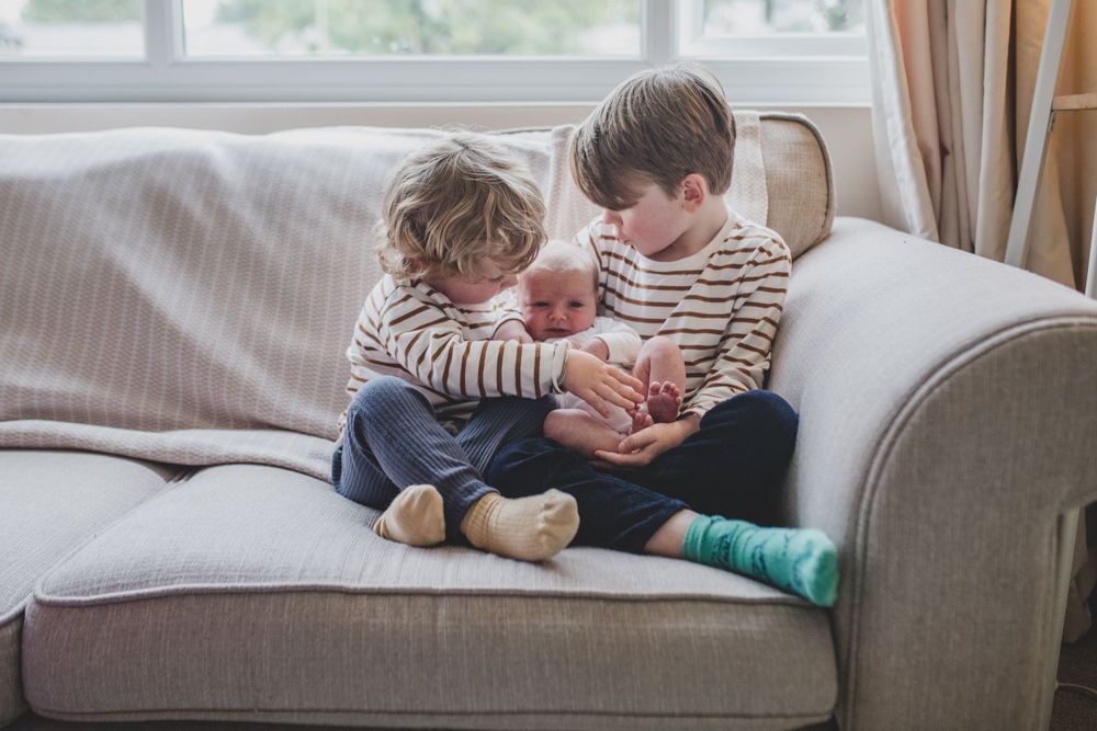 Two siblings in striped shirts sit together on a gray couch by a window sharing a tender moment.